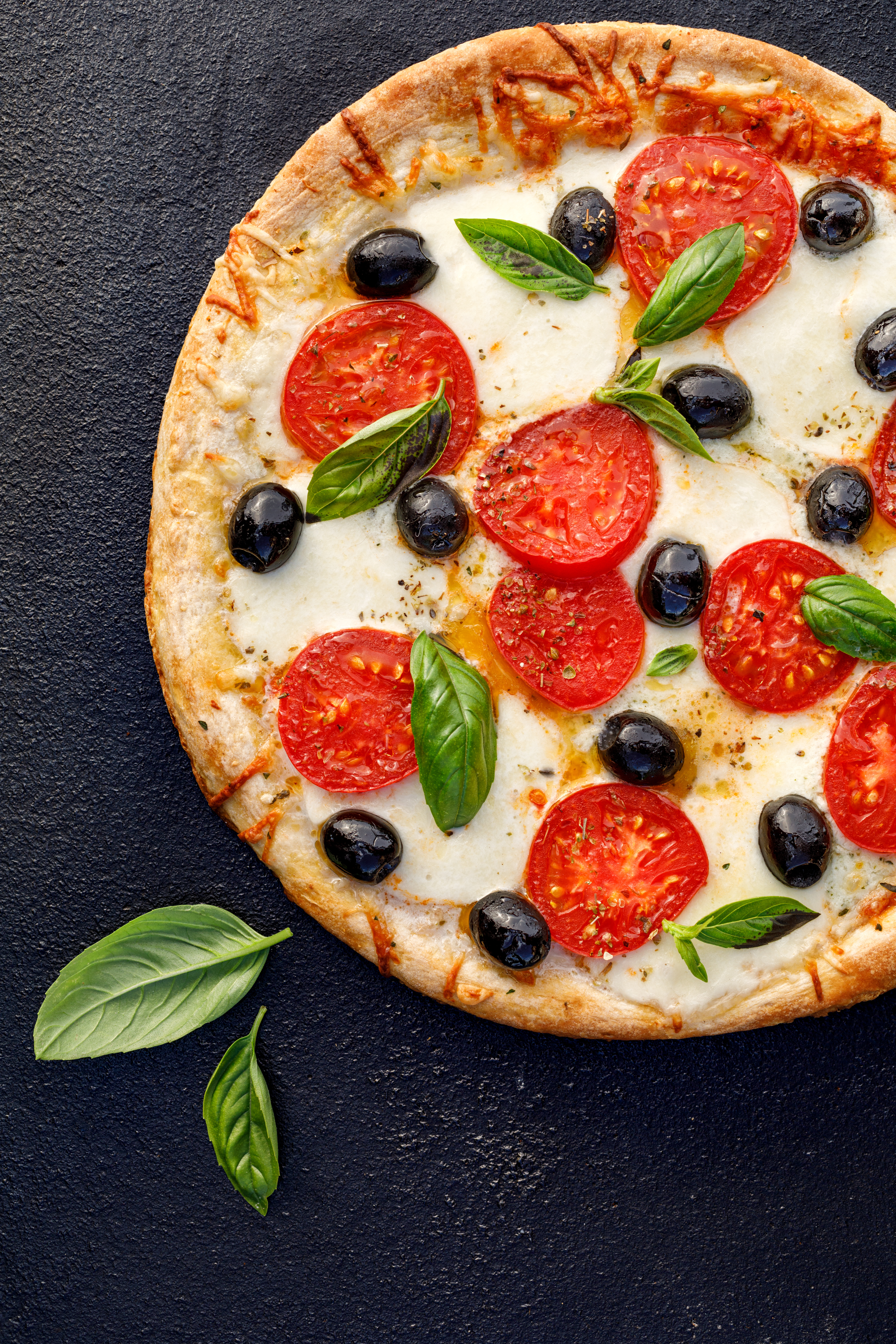 Pizza with tomatoes, mozzarella cheese, black olives and fresh basil on a black background, close-up, top view.
